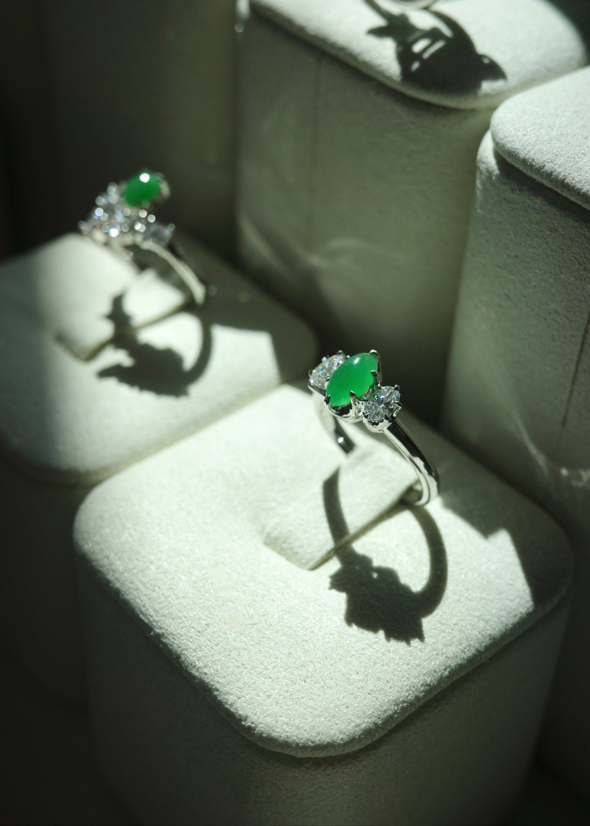 Perspective shot of two natural marquise green jade and diamond three stone rings on velvet stands, casting structured light and shadow details.