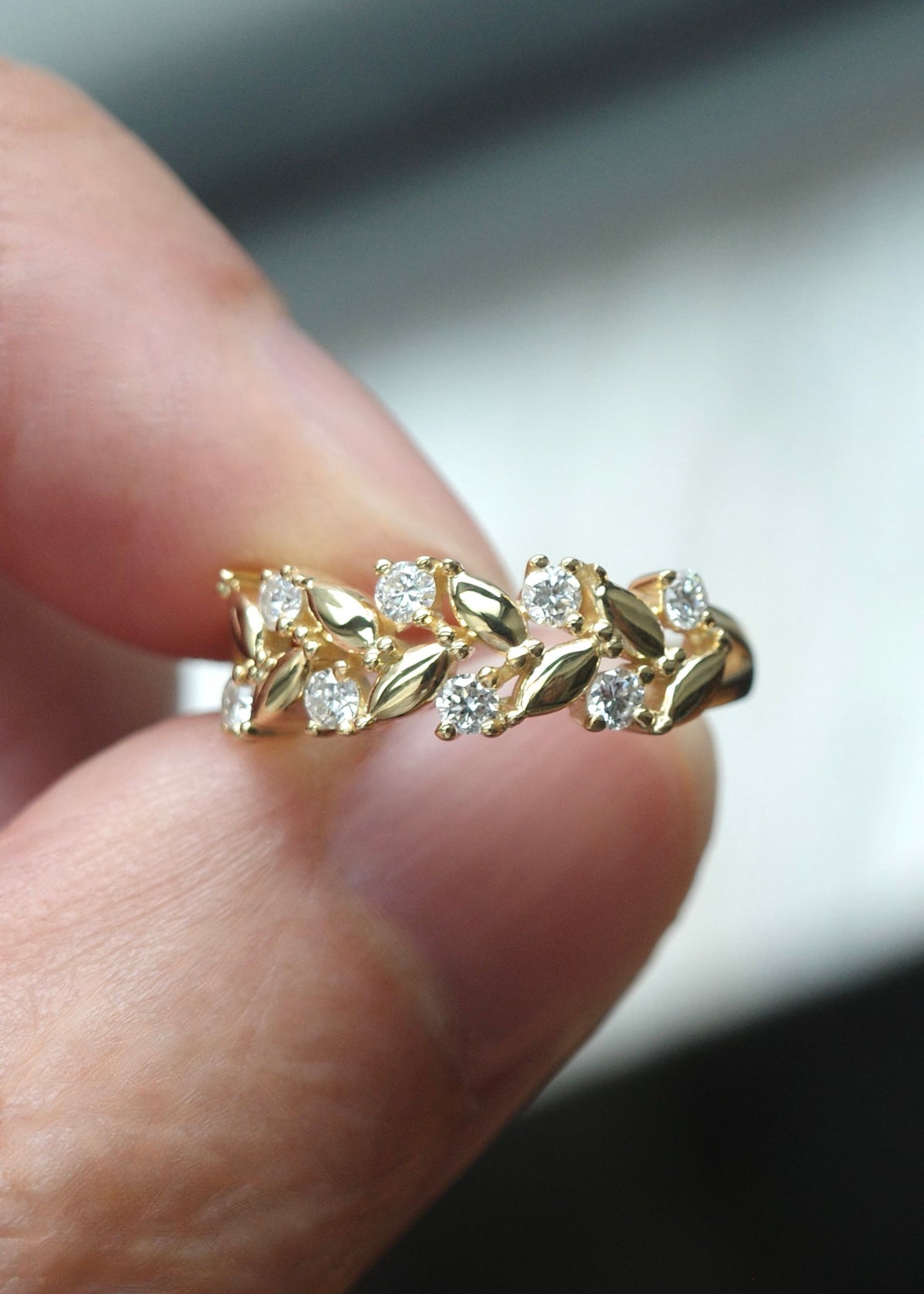 A close-up of a delicate gold ring by HN Jewelry with round brilliant cut diamond held between two fingers against a blurred gray and black background.