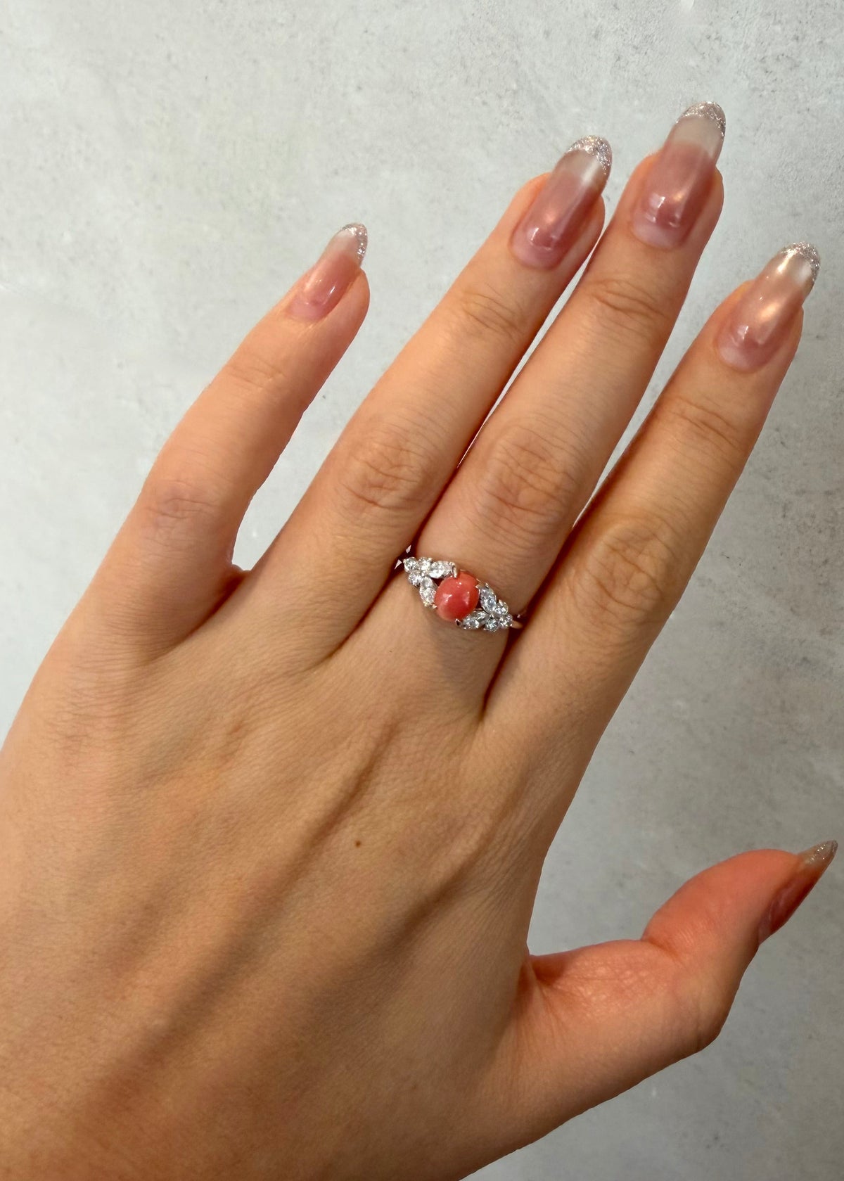 A close-up of a hand wearing a white gold ring with a central oval coral pearl and marquise diamond, set against a light gray textured background.