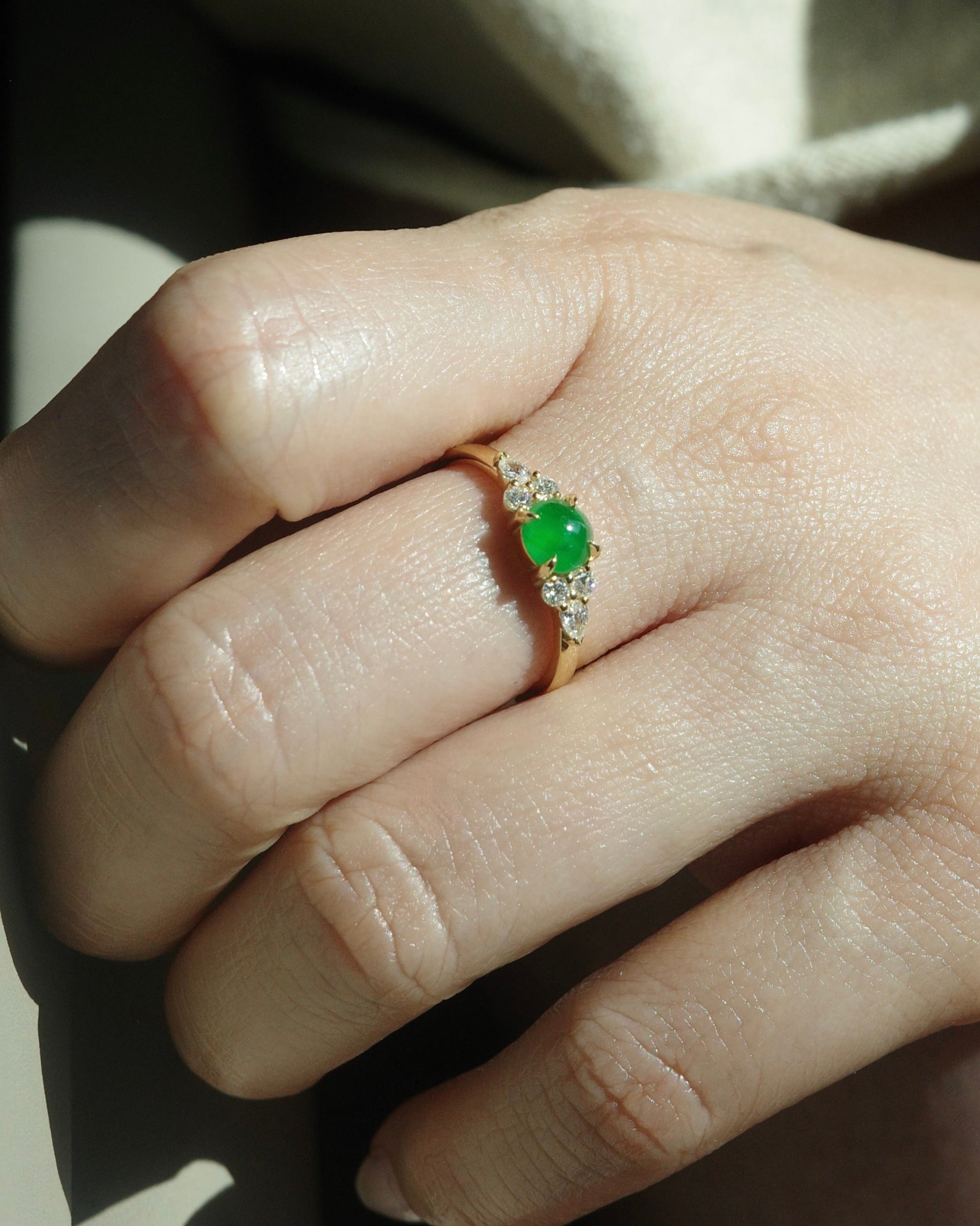 Hand wearing a ring with a 5mm round jadeite at the center along with pear diamonds and round brilliant cut diamonds on the side, made in 14k solid yellow gold