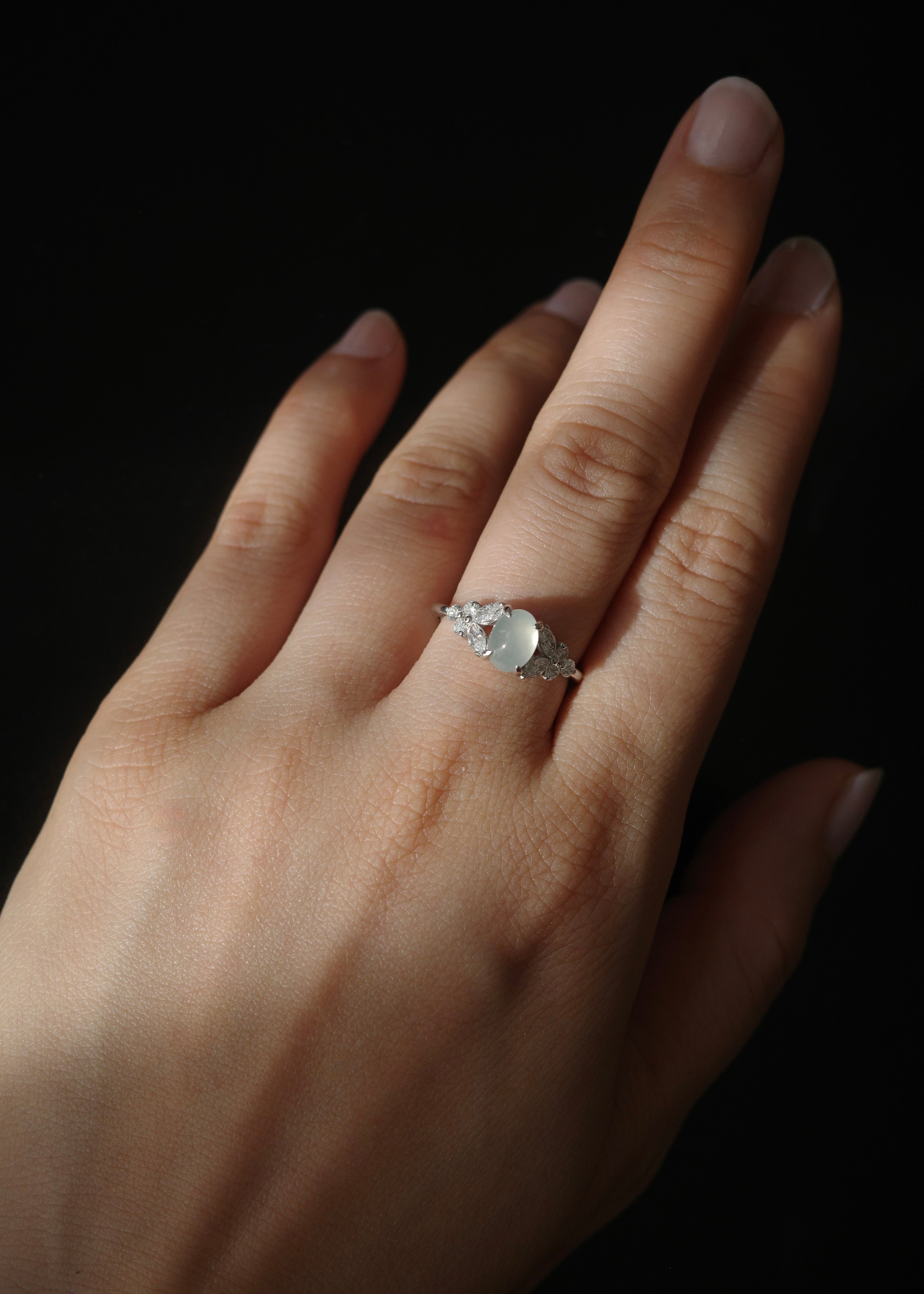 Hand wearing a white jade ring with a marquise diamond and round diamond on a dark background