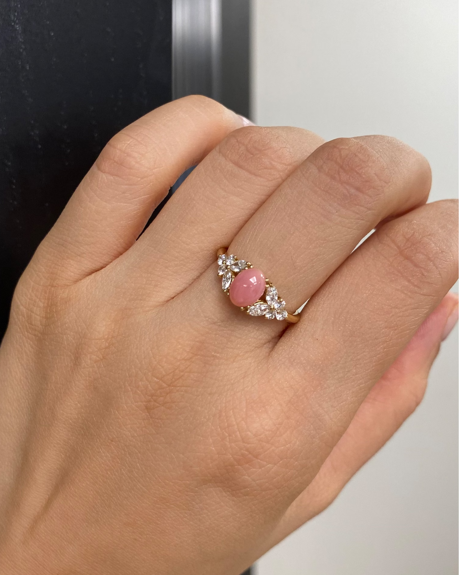 Hand wearing a ring with a pink gemstone and diamond accents against a neutral background