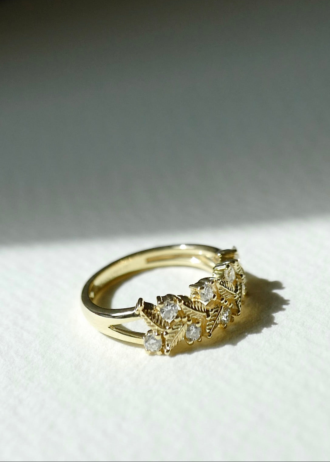 A gold ring with delicate leaf and vine motifs set with small clear stones, resting on a textured white surface under dramatic side lighting.
