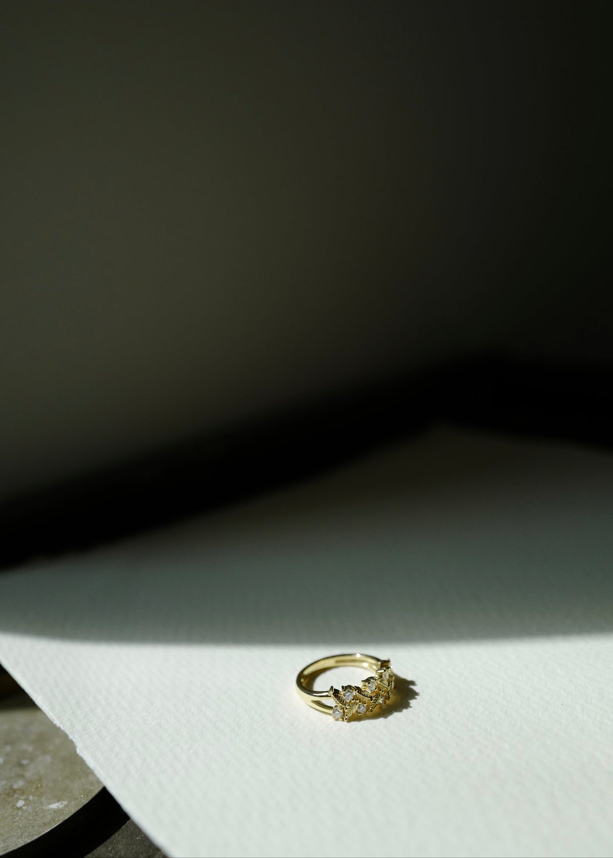 A delicate gold ring with a small floral or leaf-like cluster setting, resting on a textured white surface under dramatic, high-contrast directional lighting.