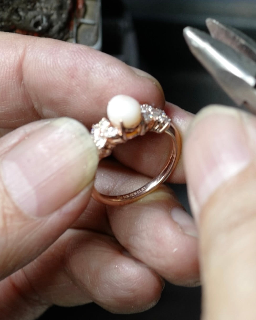 A close-up of a jeweler hand holding a rose gold ring with an oval white oval conch pearl and marquise diamonds.