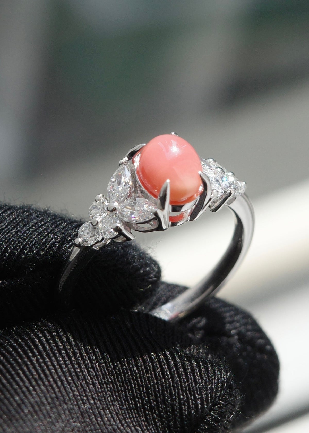 A white gold ring with a oval coral pearl center stone and diamond accents, held on a black textured glove against a blurred background.