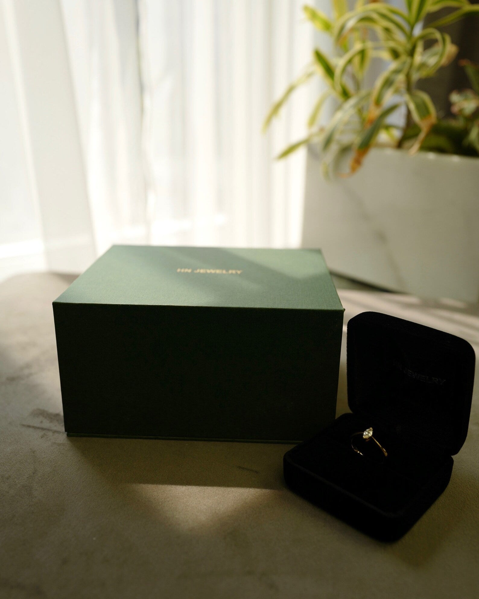 Black jewelry box with a ring on a light surface next to a green box and plant.