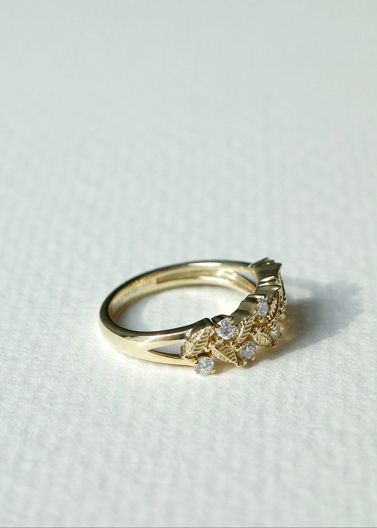 A gold ring with a delicate filigree band and a cluster of diamonds on the front, photographed on a textured off-white surface.