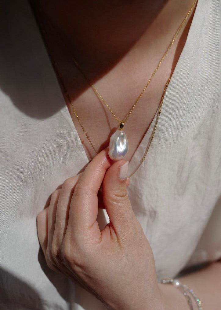 Hand holding a pearl necklace with a pearl pendant against a neutral background