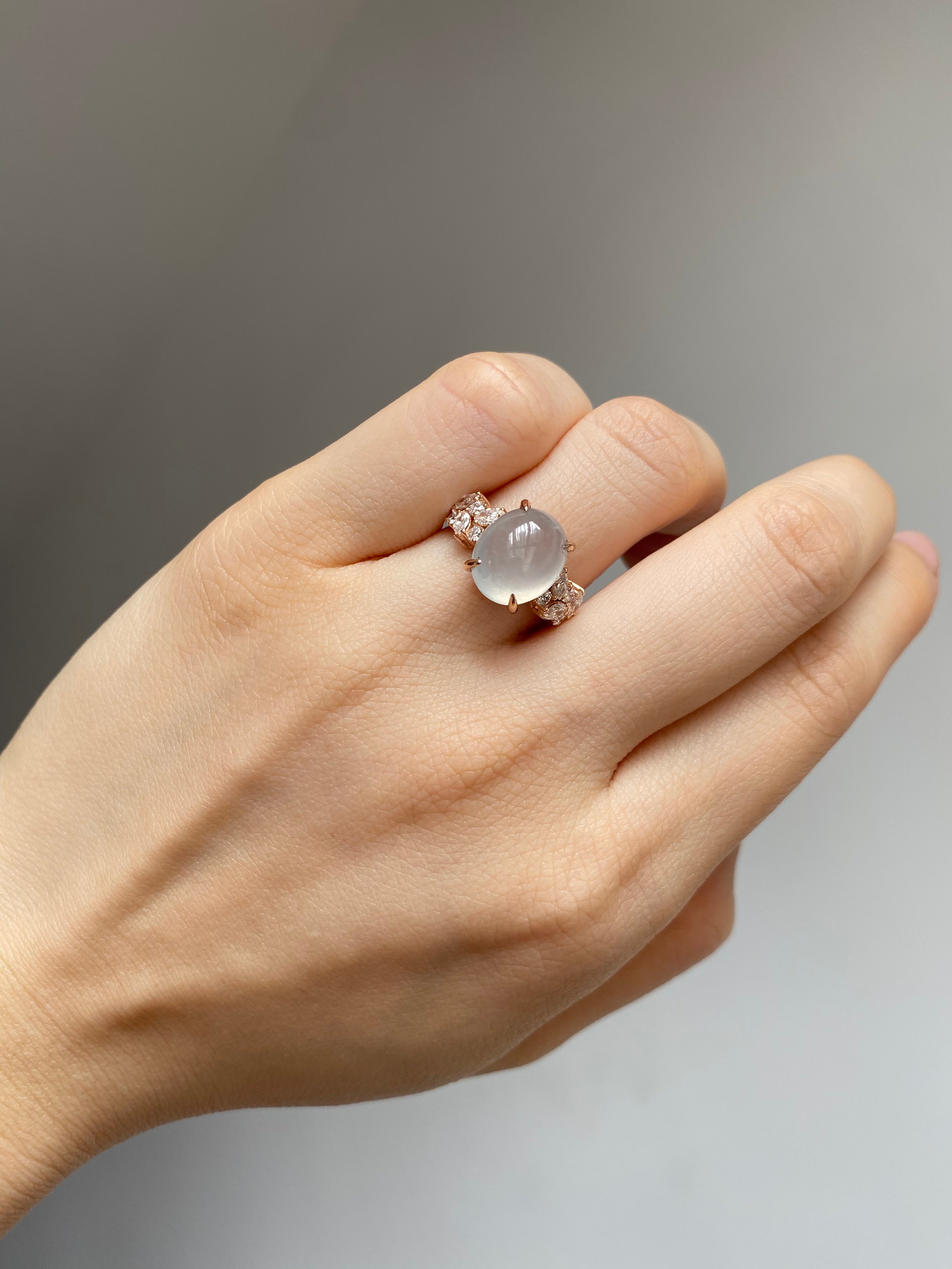 Hand wearing a ring with a natural oval icy white jade at the center and marquise diamonds on the side in 14k solid rose gold