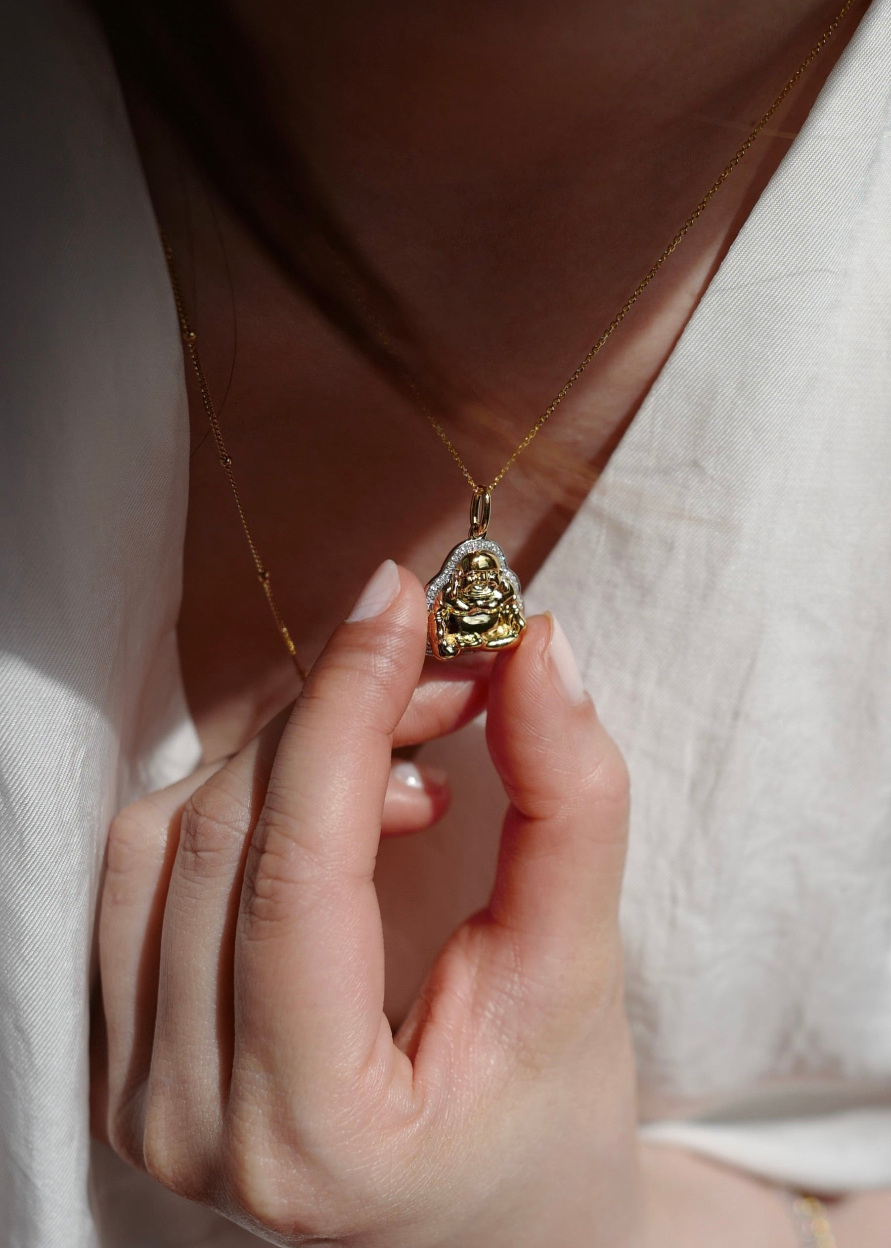 Hand holding a gold necklace with a Buddha pendant against a neutral background