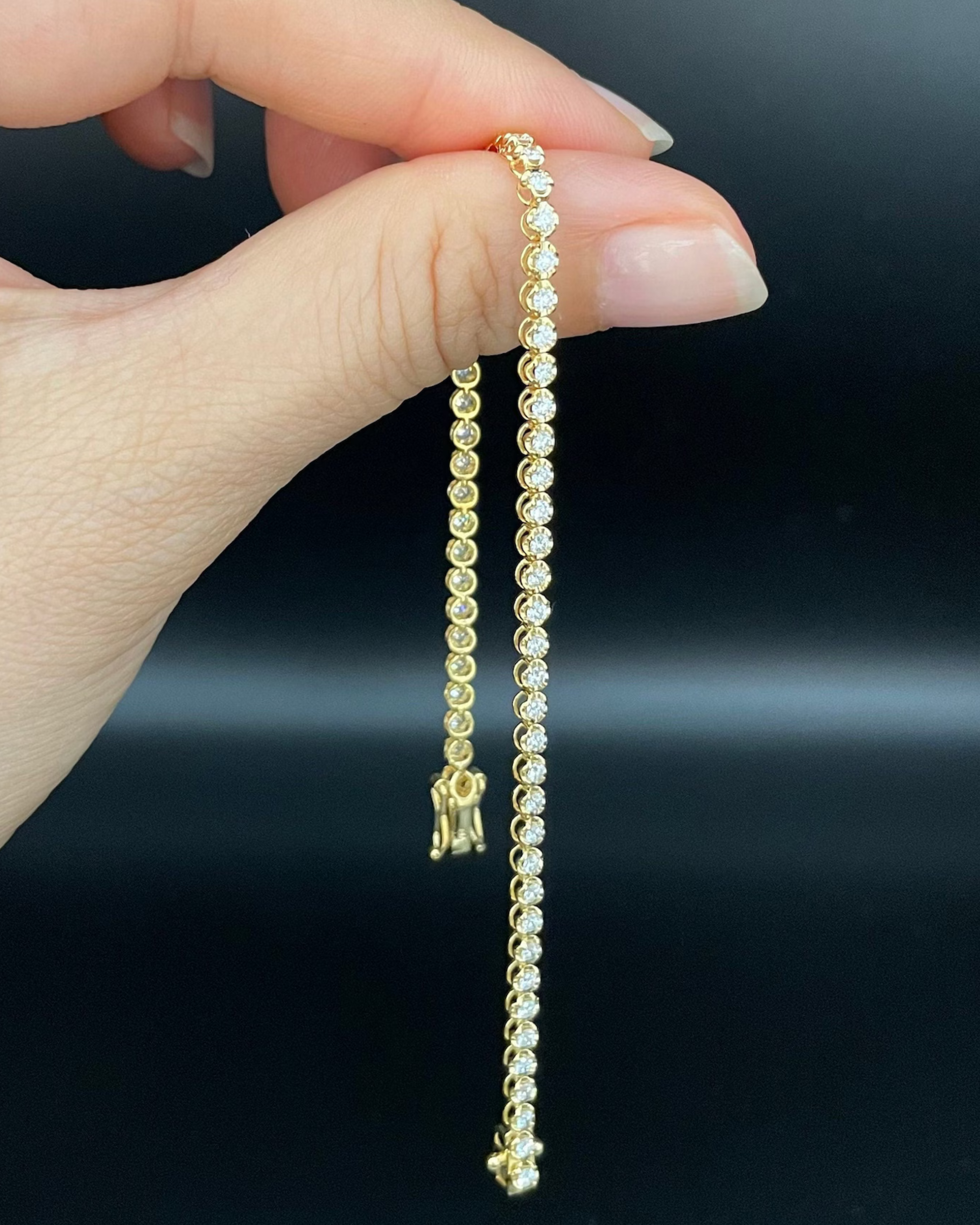 Tennis bracelet with round brilliant cut diamonds made in yellow gold held by a hand against a dark background