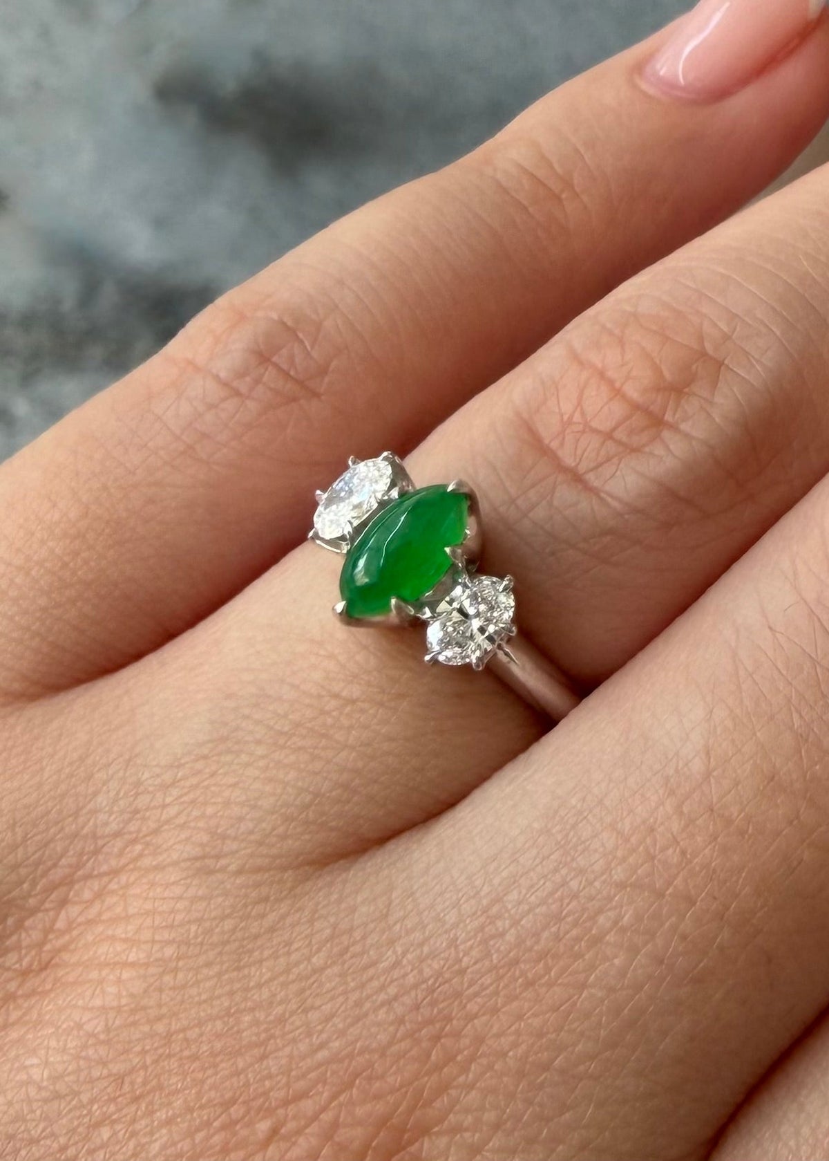 Close-up view of a natural green jade marquise cabochon ring on hand, featuring two brilliant natural oval diamond accents in solid white gold prongs.
