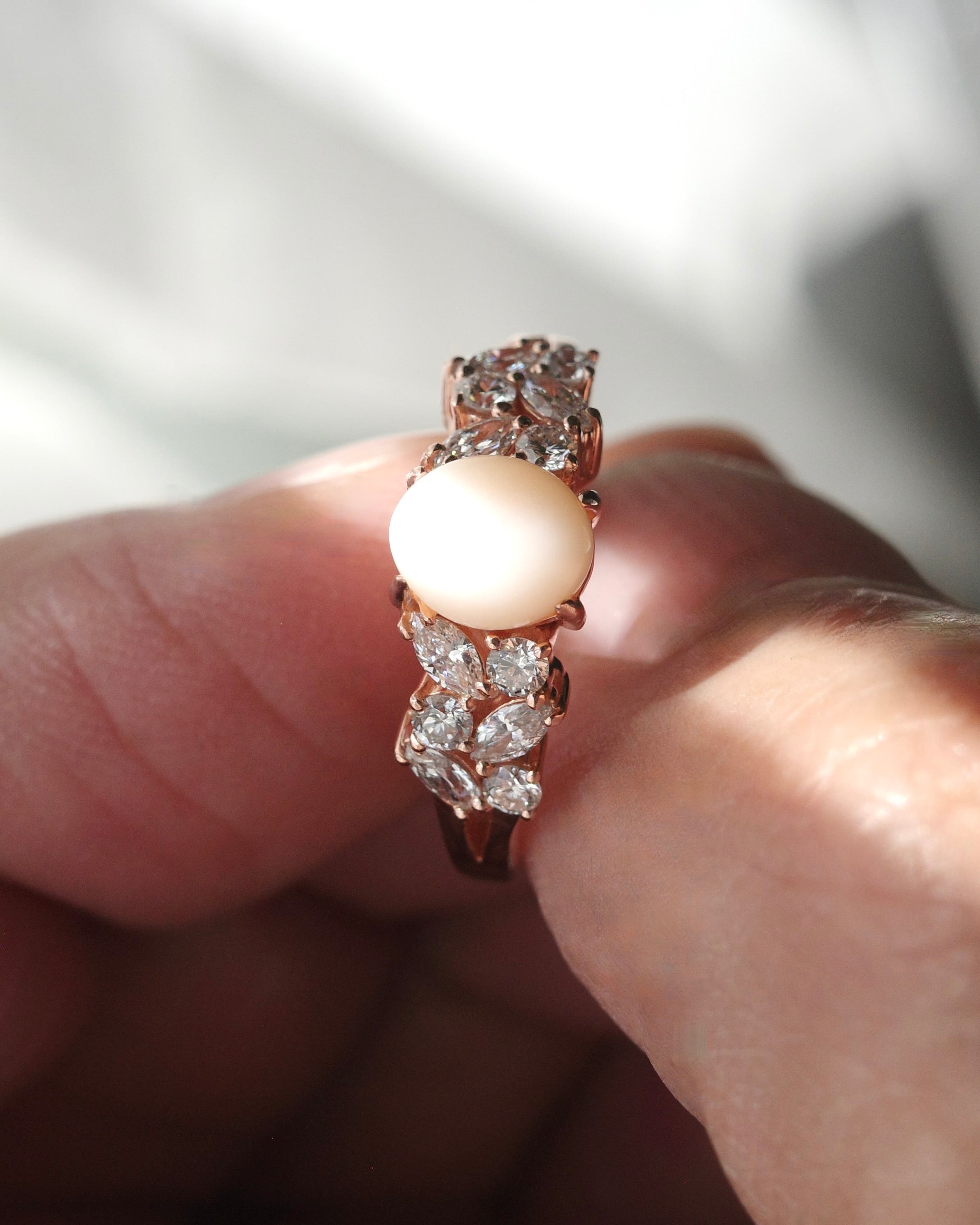 A rose gold ring with a oval white pink conch pearl at the center along with diamond accents, held between fingers.