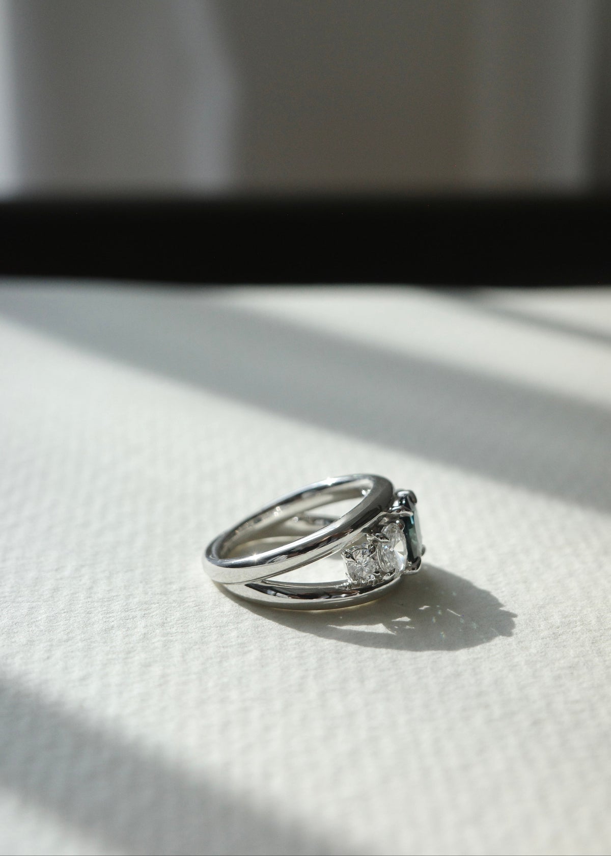 Natural oval teal sapphire five-stone ring set in solid 14k white gold on white textured paper by HN Jewelry.