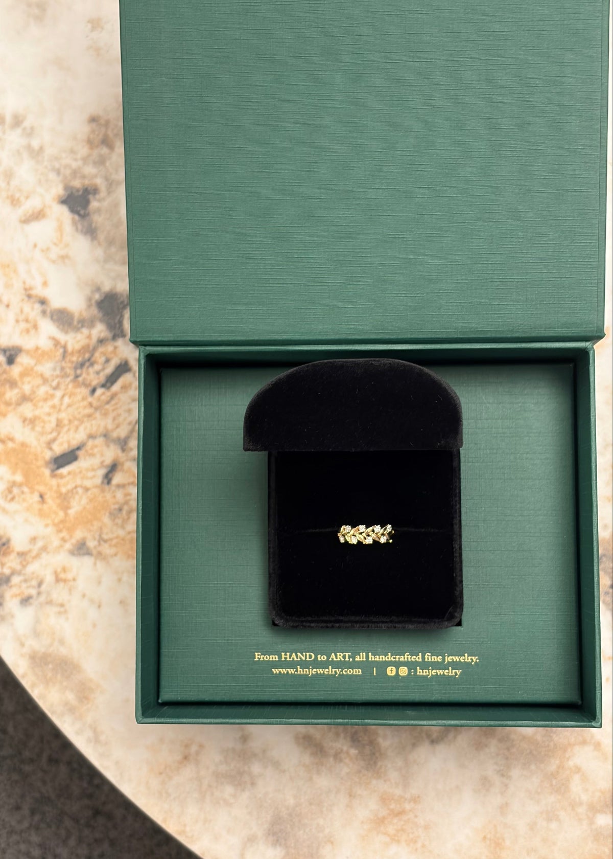 A gold ring with small diamonds in a black velvet HN Jewelry ring box, presented inside an open textured green gift box on a marble surface.