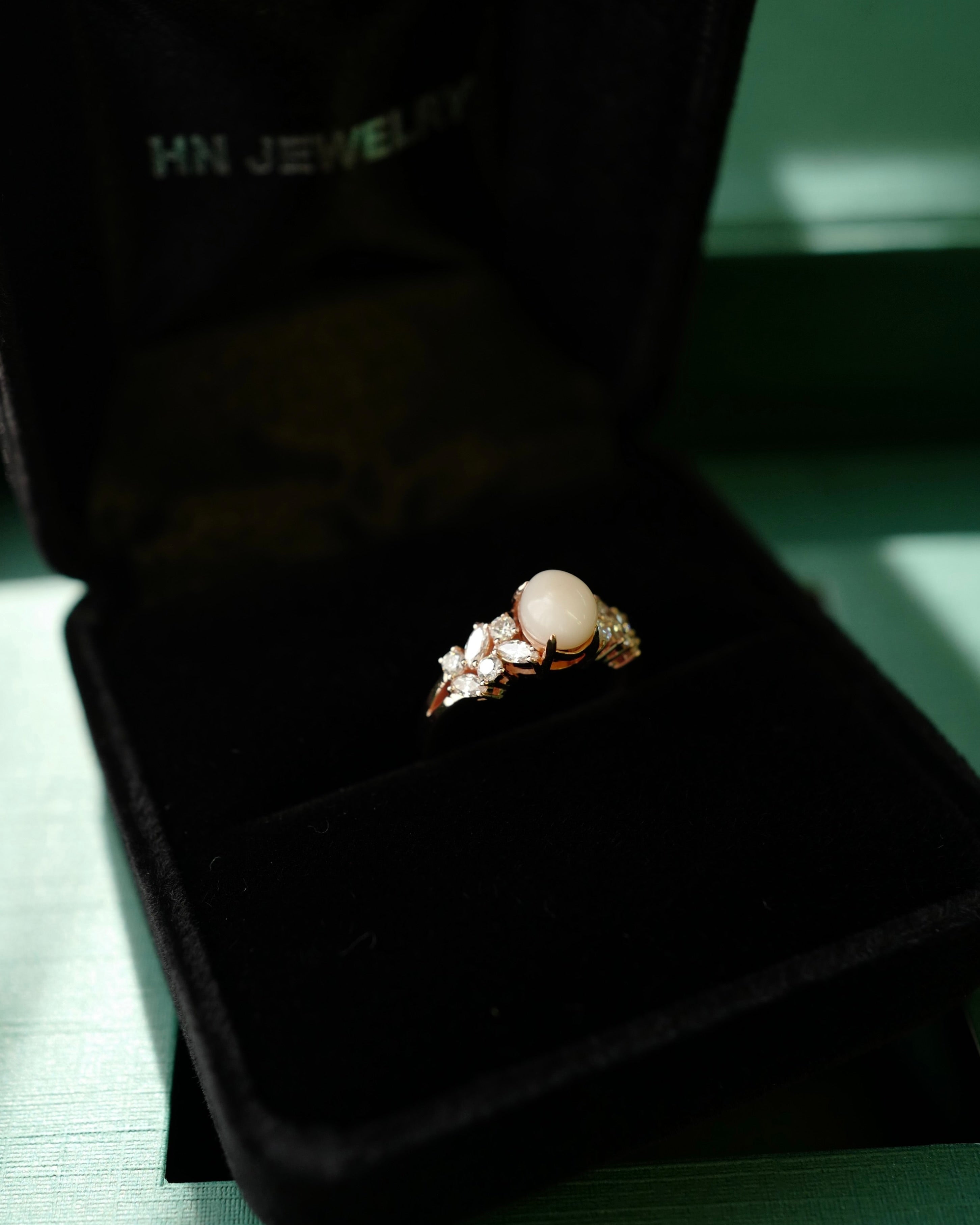 A rose gold ring set with an oval white conch pearl and marquise diamond and round diamonds displayed in an open black HN Jewelry package box.