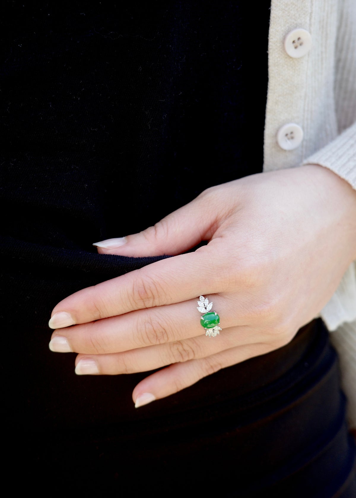 A close-up of a person's hand wearing a white ring with a green jade center stone and small marquise side stones, styled with a black top and cream cardigan.