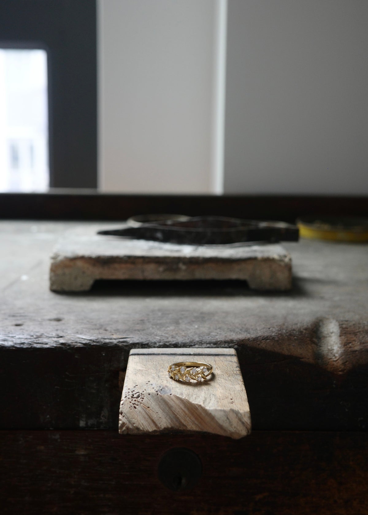 A gold ring with a detailed filigree or floral design, displayed on a small wooden block on a rustic dark wooden workbench.