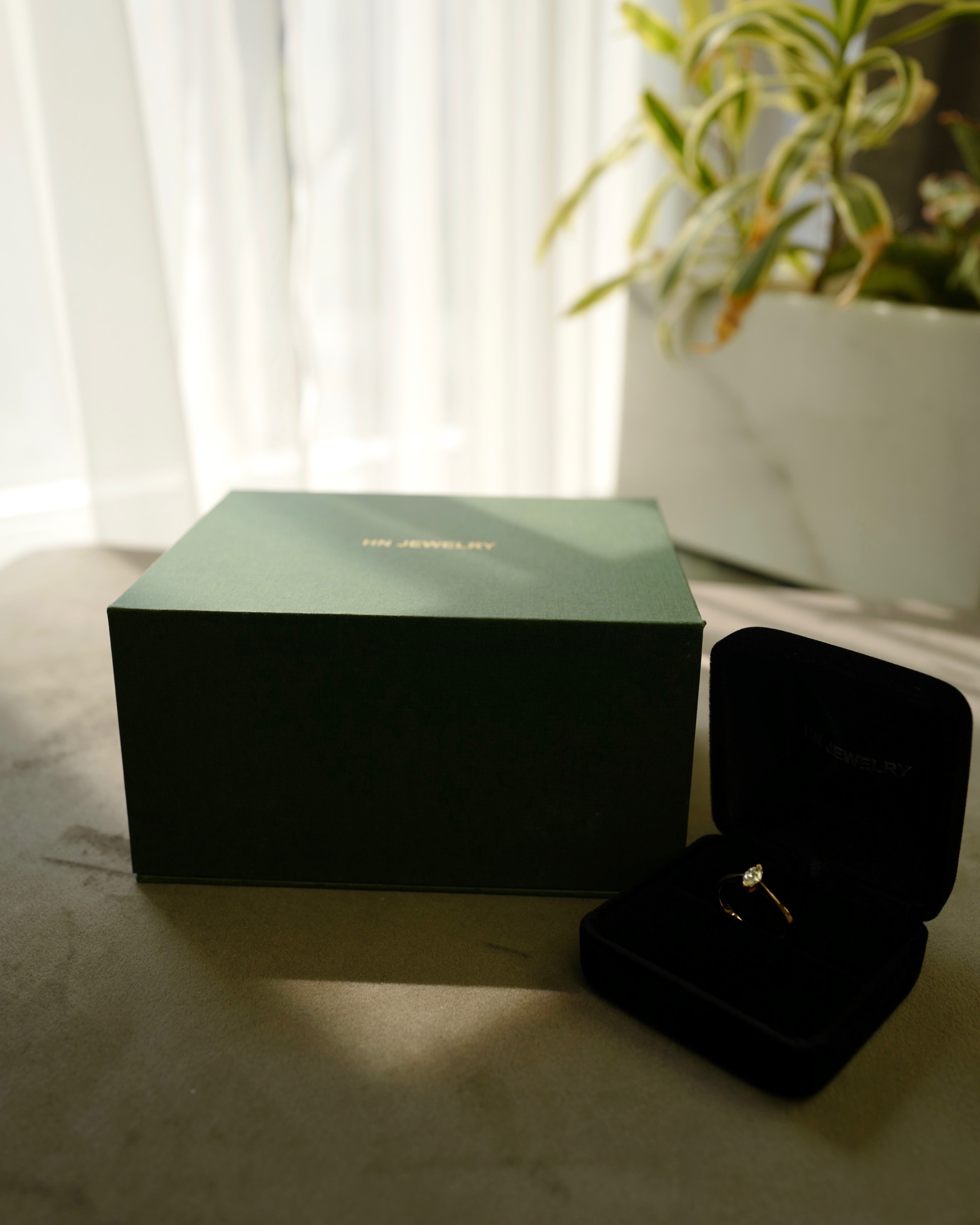 Black jewelry box with a ring on a light surface next to a green box and plant.