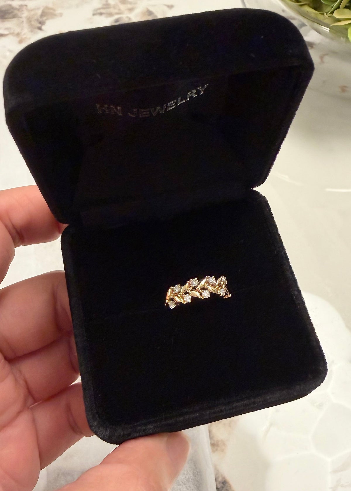 A gold ring with small diamonds set in a leaf-like pattern, displayed inside an open black velvet HN Jewelry jewelry box held by a hand.
