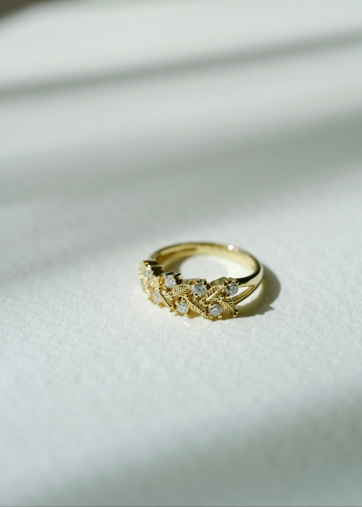 A gold ring with a textured, leaf-like or vine-like design set with small diamond, resting on a white textured surface with soft diagonal shadows.