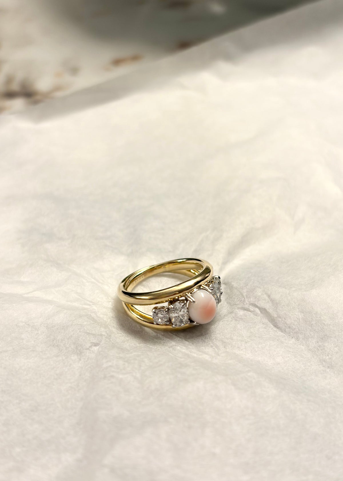 A gold ring featuring a central round pale pink conch pearl and two diamonds on either side, resting on textured white paper.