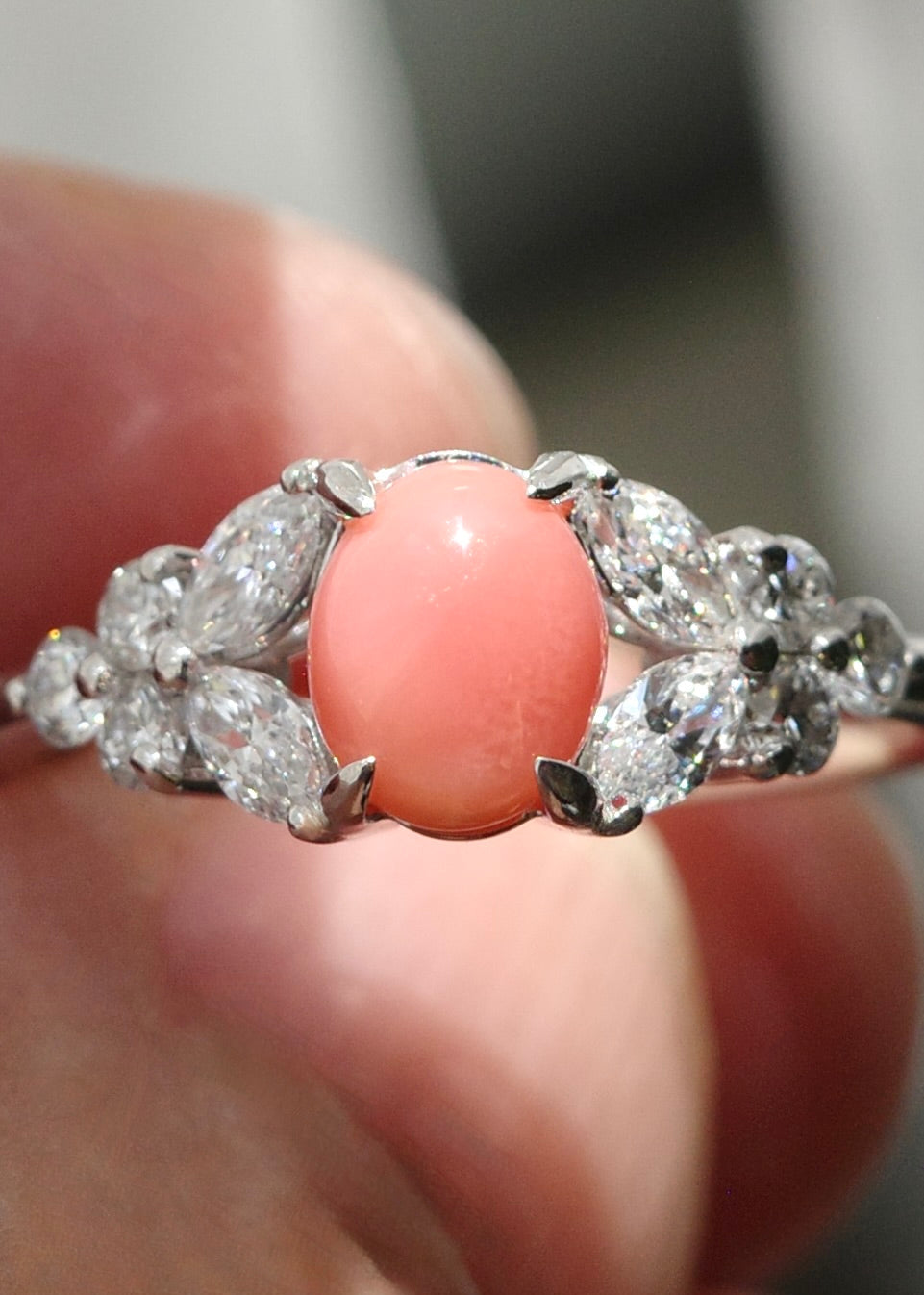 A close-up of a ring held on fingers, featuring a smooth oval coral pearl at center surrounded by marquise and round diamond in a white gold setting.