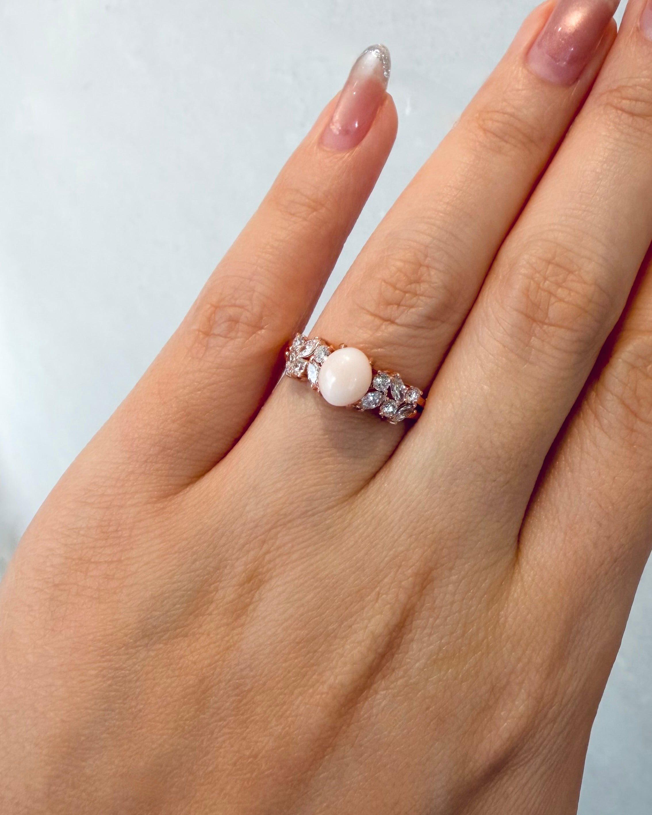 A close-up of a hand wearing a rose gold ring with a central oval white conch pearl and high quality marquise diamonnd accent on the side, set against a light gray background.