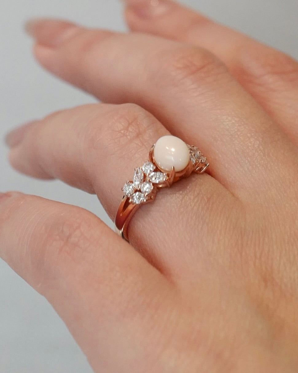 A close-up of a hand wearing a rose gold ring with a central oval white pink natural conch pearl and marquise diamond and round brilliant cut diamond accents on either side of the band.